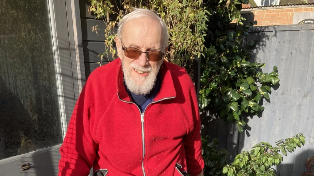 Tony wears a red fleece and smiles at the camera while standing in his garden