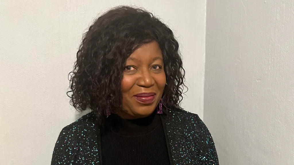 A woman stands in front of a white wall and smiles at the camera. She is wearing a sparkly black cardigan and black turtleneck.