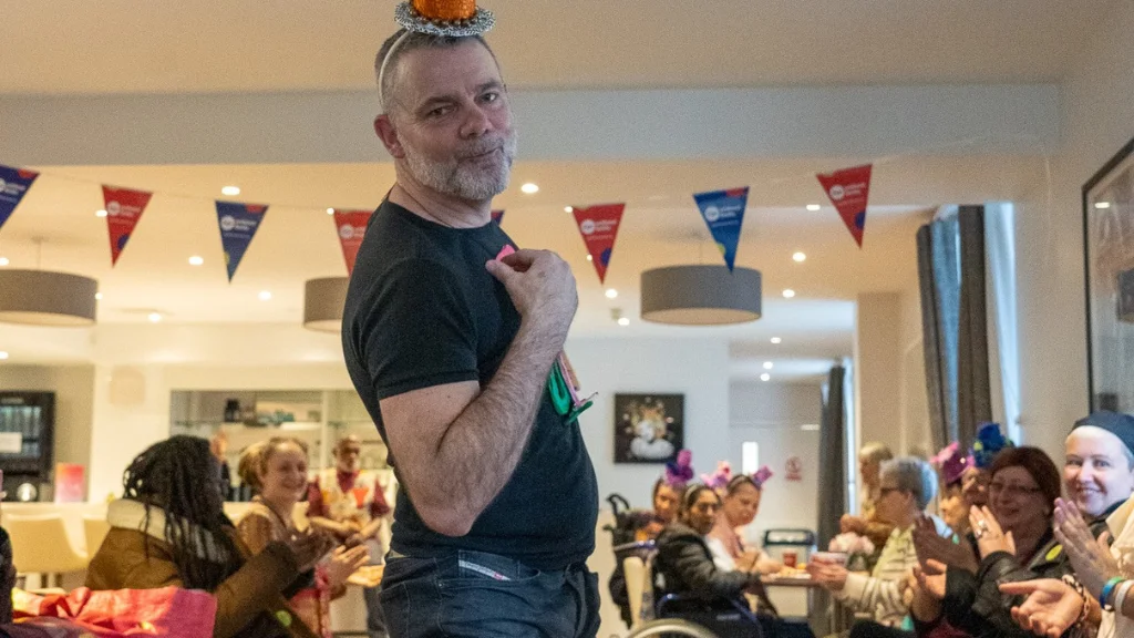 A man wearing an orange and green hat stands and poses for the camera. In the background there are people sitting and clapping and Age Without Limits bunting is overhead.