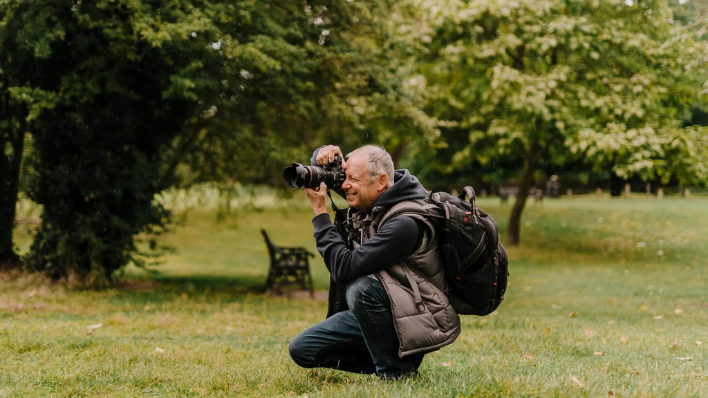 A man crouches down on grass and takes a photo using a camera.