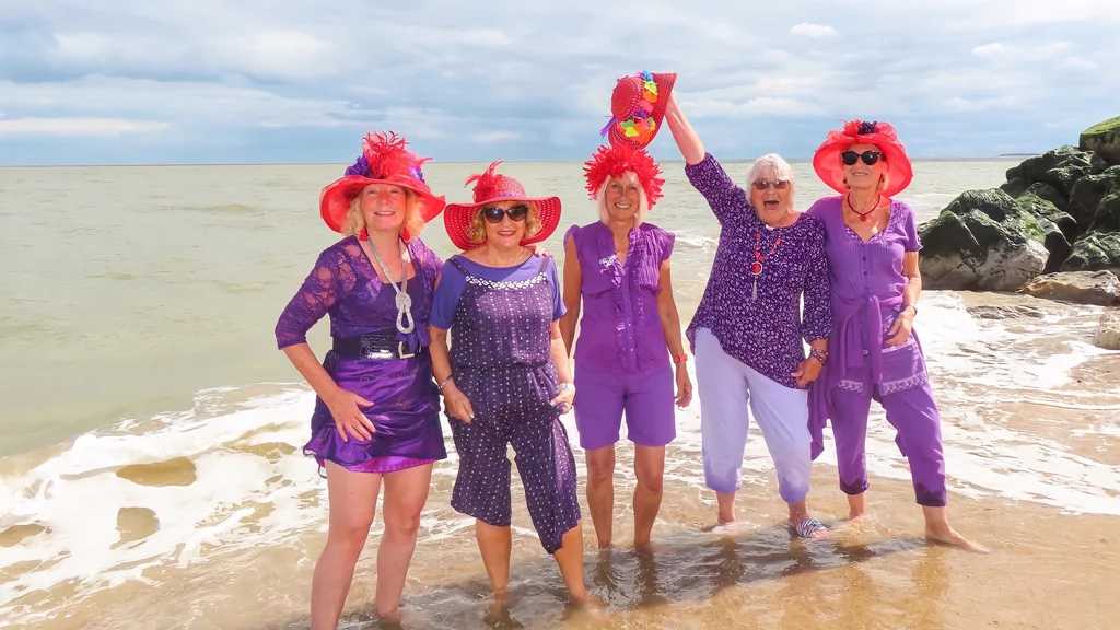 A group of older women dressed in purple with red hats stand at the sea line on a beach.