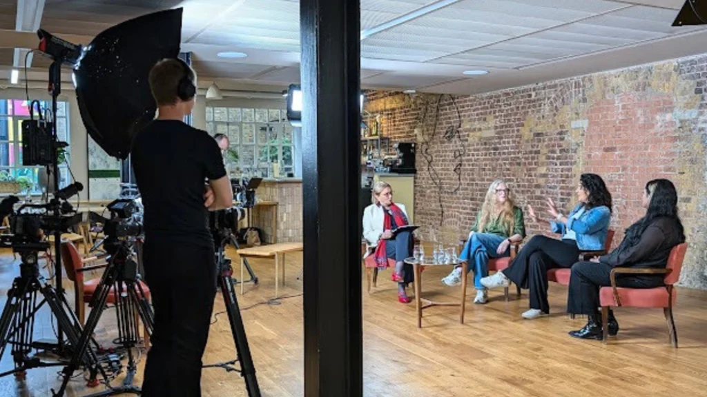 A behind the scenes image showing four women sat on chairs in a row with a film crew in front of them.