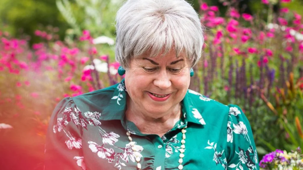Pauline wears a green floral dress and looks downwards, smiling, with colourful flowers in a garden behind her.