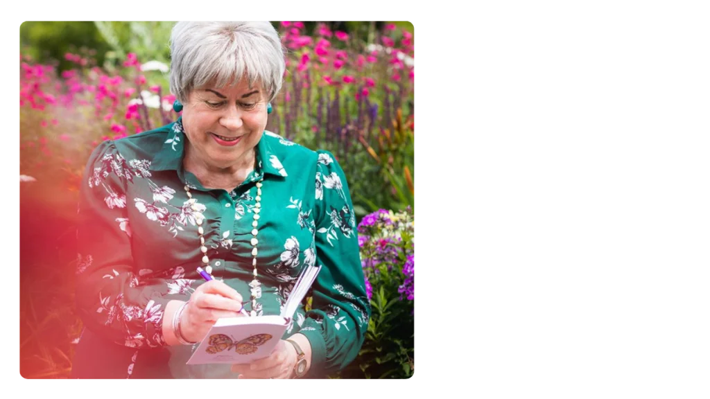 A woman wearing a green floral dress smiles and writes in a notebook, with a colourful garden of flowers behind her.