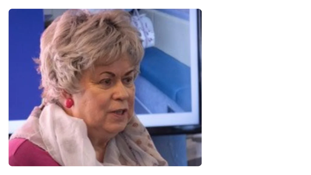 A woman wearing a pale pink scarf and pink earrings talks with a presentation behind her.